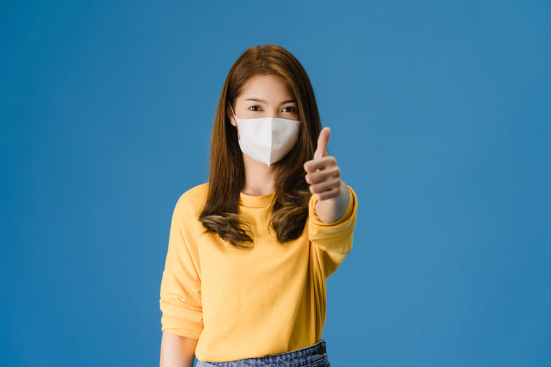 young-asia-girl-wearing-medical-face-mask-showing-thumb-up-with-dressed-casual-cloth-look-camera-isolated-blue-background-self-isolation-social-distancing-quarantine-corona-virus